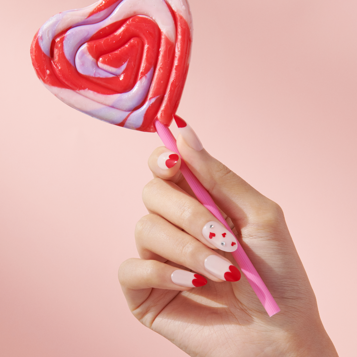 A hand with red, pink, and white heart-themed nail art holds a heart-shaped red and white swirled lollipop against a pink background.